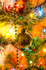 Christmas golden decoration hanging on a Christmas tree branch with bright colorful holiday lights. Background with golden Christmas ball, and bright bokeh, Christmas and New Year background in warm 