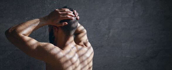 A man stands in the shower, water cascading down, with sunlight creating stripes on his bare back....