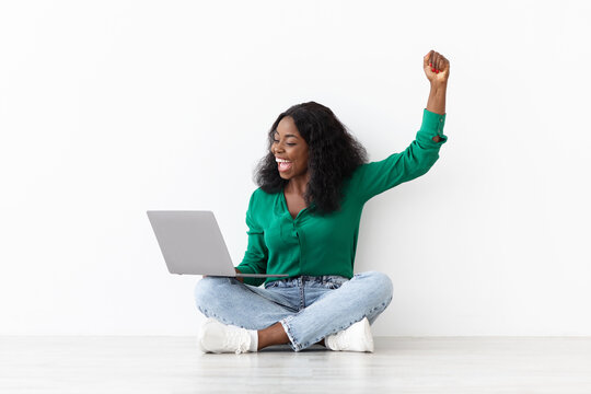 A woman with curly hair sits cross-legged on the floor, celebrating happily as she interacts with her laptop in a well-lit room during the day.