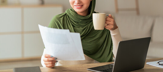 A young woman sits comfortably at a desk with a laptop, holding a cup and reviewing papers. She is dressed in a green scarf, enjoying her cozy workspace and focusing on her tasks.