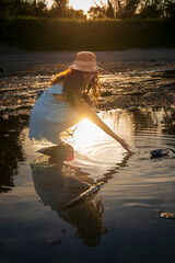 Woman playing with paper boat in water reflecting warm sunset colors