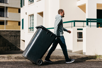 Man in casual attire walking with a black trash bin along a residential street, showcasing urban living and community responsibility in a clean environment