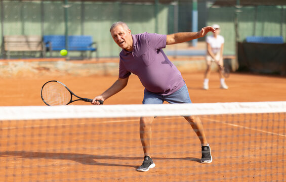 Elderly couple in sportswear spending time playing sports on tennis courts on sunny day, old man receiving hit from tennis racket - Powered by Adobe