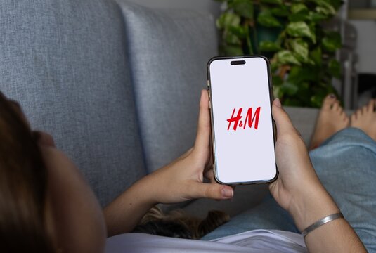 Rosario, Argentine. November,18,2025:Cute young woman relaxing on a couch at home, using a smartphone to browse an online fashion retail application, enjoying a comfortable mobile shopping experience