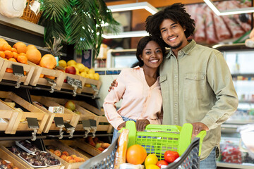 A happy couple stands close together in a lively grocery market, smiling while holding a cart filled with colorful fruits and vegetables, surrounded by fresh produce displays.
