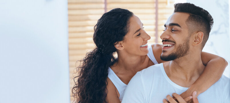 A smiling couple enjoys a happy moment together inside their home. The sunlight filters through the window, highlighting their affection and connection. They look at each other with love.