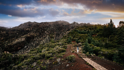 Scenic view of a couple walking along a stone path through lush greenery and rocky terrain, surrounded by dramatic clouds and mountains, capturing the essence of adventure and romance