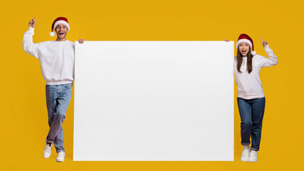 A joyful couple dressed in festive attire smiles as they hold a large blank sign with one hand raised in excitement. The bright yellow backdrop adds a cheerful vibe, perfect for holiday promotion.
