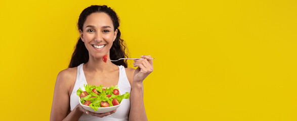 A woman with long curly hair enjoys a healthy salad in a bright setting. She takes a bite of lettuce and shows enthusiasm for her meal. The yellow background adds vibrancy.