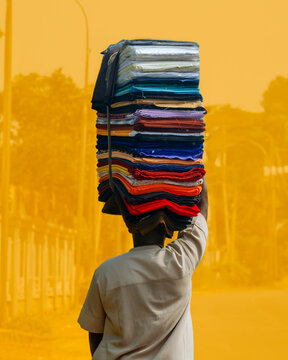 View of a figure silhouetted against a vibrant yellow backdrop, balancing a towering stack of colorful fabrics in old gra, Port Harcourt, Rivers, Nigeria.