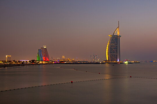 View of the iconic Burj Al Arab and other illuminated buildings sparkle against the tranquil waters at dusk, Dubai, Dubai, United Arab Emirates.