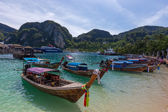 View of longtail boats resting on the turquoise waters before a backdrop of lush green cliffs under a bright blue sky, Phi Phi Island, Krabi, Thailand.