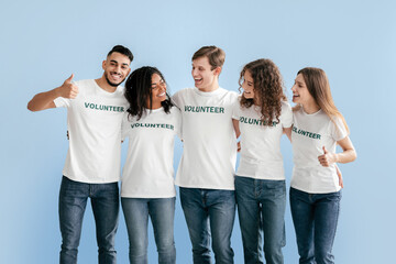 Five young adults stand closely together, wearing matching shirts that say Volunteer. They smile and engage with each other, celebrating teamwork and community spirit in a bright, cheerful setting.