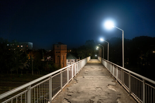 Night view of an urban pedestrian overpass with bright streetlights and clean geometric lines. Modern city infrastructure, minimalistic architecture and clear night sky create a calm, futuristic atmos