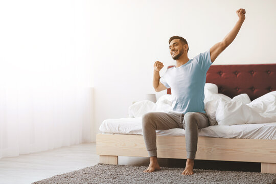A young man sits on the edge of his bed, stretching his arms and smiling as sunlight fills the cozy bedroom. The room has white walls and a neatly made bed with fluffy pillows.