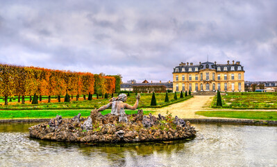 The Bassin de Scylla fountain in the foreground of the Chateau de Champs-sur-Marne near Paris. The historic French castle and formal gardens are seen under a cloudy sky