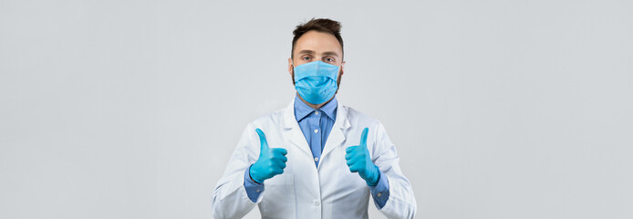 A healthcare professional stands confidently with a thumbs-up gesture. He is wearing a blue mask and gloves, conveying a message of safety and reassurance in a neutral environment.