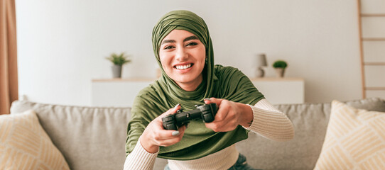 A young woman wearing a green hijab is smiling while playing video games at home. She holds a gaming controller and is seated on a couch in a bright living room.