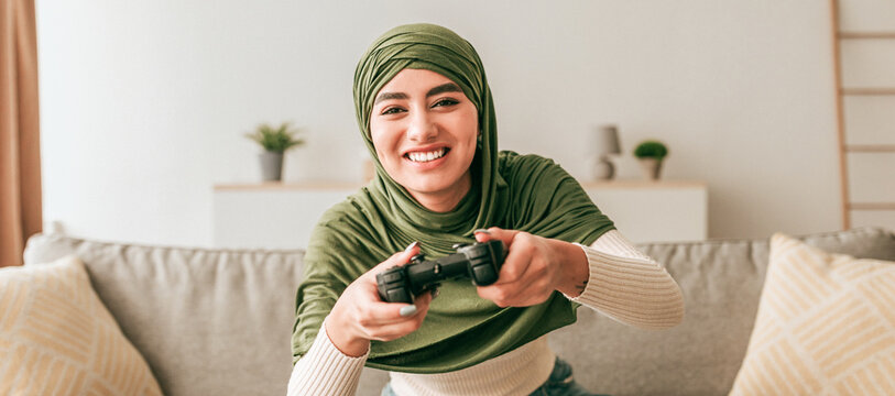 A young woman wearing a green hijab is smiling while playing video games at home. She holds a gaming controller and is seated on a couch in a bright living room.