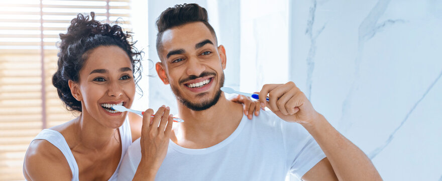 A couple is happily brushing their teeth together in a bright, modern bathroom. They smile at each other while keeping a playful atmosphere, showcasing a cheerful morning moment.