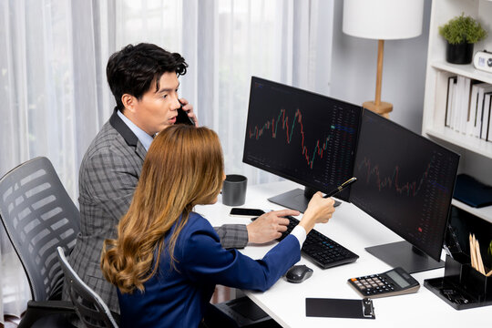 Smiling Asian businessman calling to broker dynamic stock exchange database with woman business partner pointing to screens comparing profit value online application program at home office. Infobahn. - Powered by Adobe