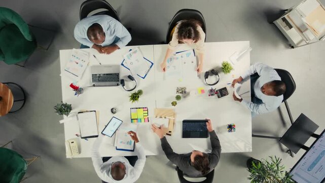 Top down view zoom in of diverse colleagues and managers participating in briefing. Business meeting focuses on negotiation, reporting and resource allocation for development and success. Camera A.