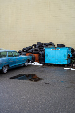 Old Blue Car Parked Near a Pile of Tires and a Dumpster