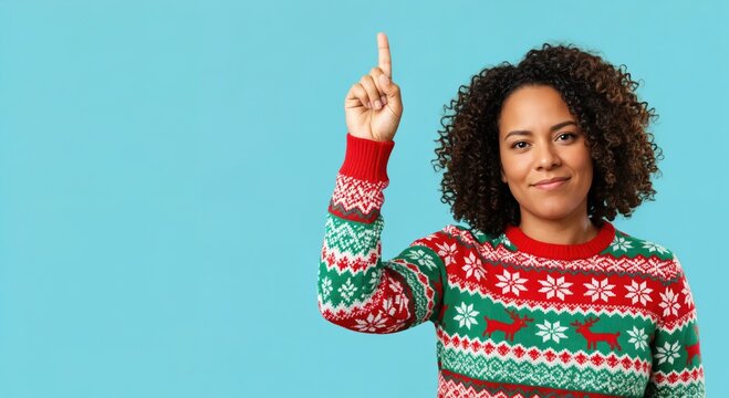 Smiling young African American woman in a festive red and green Christmas sweater pointing her index finger upwards, isolated on a blue background with copy space.
