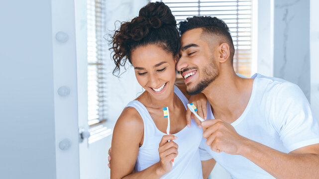 A young couple smiles at each other while brushing their teeth in a well-lit bathroom. The atmosphere is cheerful and playful, showcasing their shared morning routine and connection.