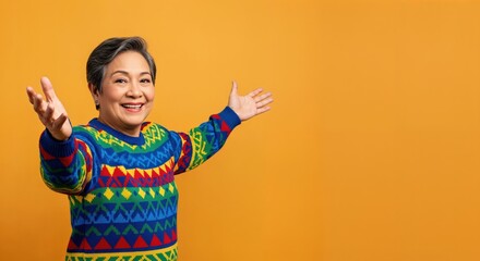 Joyful elderly Asian woman with short grey hair wearing a colorful patterned sweater, welcoming with open arms against an orange backdrop.