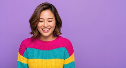 Fototapeta premium Joyful young Asian woman with a radiant smile and closed eyes, wearing a bright multi-colored striped sweater on a vibrant purple background.