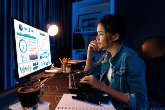 Smiling young beautiful asian creative looking on pc of profit of sales target report and writing on notebook for memo with planning invest new strategy market project at neon light night. Stratagem.