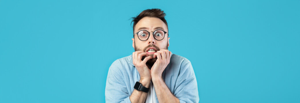 A young man with glasses and a beard displays a shocked expression. His hands are on his face, and he seems startled or frightened in a bright blue setting.