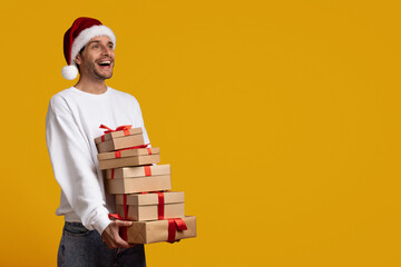 Fototapeta premium A man wearing a Santa hat smiles while carrying stacks of Christmas presents tied with red ribbons. The bright yellow background adds to the festive atmosphere of the holiday season.