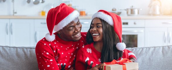 A joyful couple sits on a couch, smiling and sharing a moment together during Christmas. They wear...