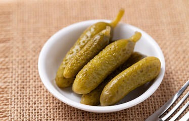 Gherkin pickle or pickled cucumber in a bowl with fork on brown background