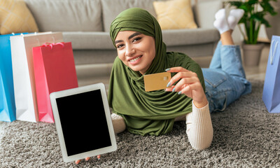 A woman wearing a green hijab relaxes on a cozy rug surrounded by colorful shopping bags. She holds a credit card in one hand and a tablet in the other, focused on her online shopping.