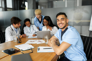 Handsome arabic man physician smiling at camera while having morning session with colleagues, multiracial group of professional doctors discussing medical cases, using modern technologies