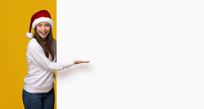 A young woman wearing a Santa hat is smiling and gesturing toward a blank space on a colorful background. She appears enthusiastic about a holiday event or promotion, embodying festive spirit.