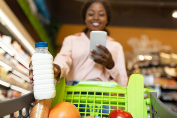 A person is holding a bottle of milk and looking at their phone while pushing a shopping cart...
