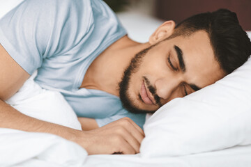 A young man is peacefully sleeping on a soft bed, surrounded by white bedding. The room has warm...