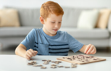 Redhead preschool boy plays alone at home, collecting letters from a wooden educational game. This...