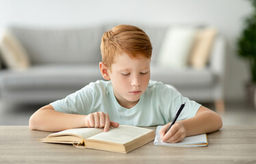 In a bright room, a smart redhead boy is focused on his homework. He is reading a book and taking...