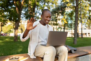 A young man sits on a bench in a park, waving at his laptop during a video call. He appears happy...