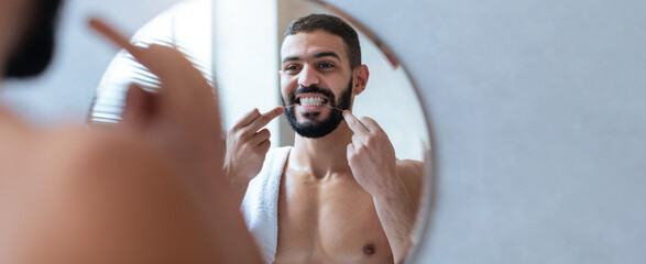 A young man stands in front of a round mirror, smiling while flossing his teeth. The bathroom is...