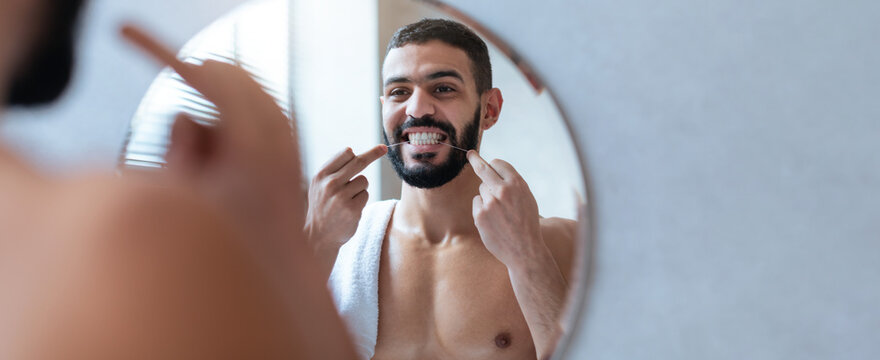 A young man stands in front of a round mirror, smiling while flossing his teeth. The bathroom is well-lit, and he is shirtless, focusing on dental hygiene. - Powered by Adobe