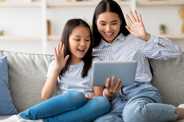 Cheerful mother and daughter are having a video call on a digital tablet. They sit on a comfortable sofa, smiling and waving at the screen. It is a joyful moment for the family.