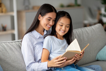 Fototapeta premium Young mother and her daughter are enjoying a cozy moment together on the sofa. They are reading a book, smiling, and sharing a bond in their comfortable living room.