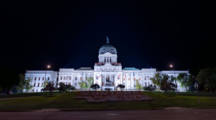 The Montana State Capitol in Helena is illuminated at night. The bright lights highlight the stone facade and dome, contrasting with the dark sky