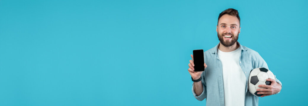 A joyful young man stands against a solid blue background holding a smartphone in one hand and a soccer ball in the other. He seems excited, likely about a recent sports update.